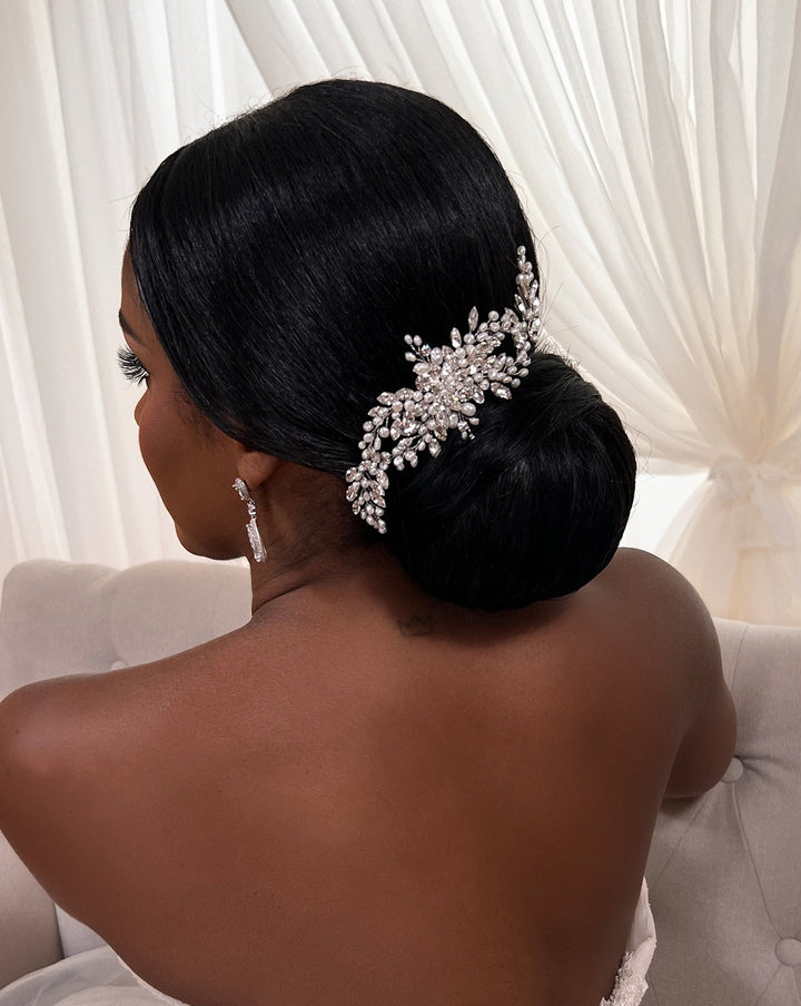A person with dark hair styled in an elegant bun is adorned with the Flowing Cluster Hair Comb, style no. 242514, featuring pearl-like details and genuine crystals. They sit on a light-colored sofa with sheer, flowing curtains in the background.