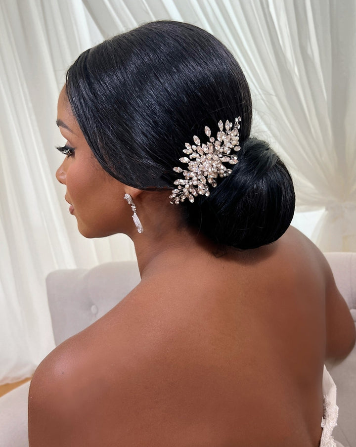 A person with sleek black hair styled in a low bun, adorned with the Dainty Swarovski Crystal Hair Comb style no. 242731, sits on a light-colored chair. They wear dangling bridal earrings and are seen from the side against a backdrop of white curtains.