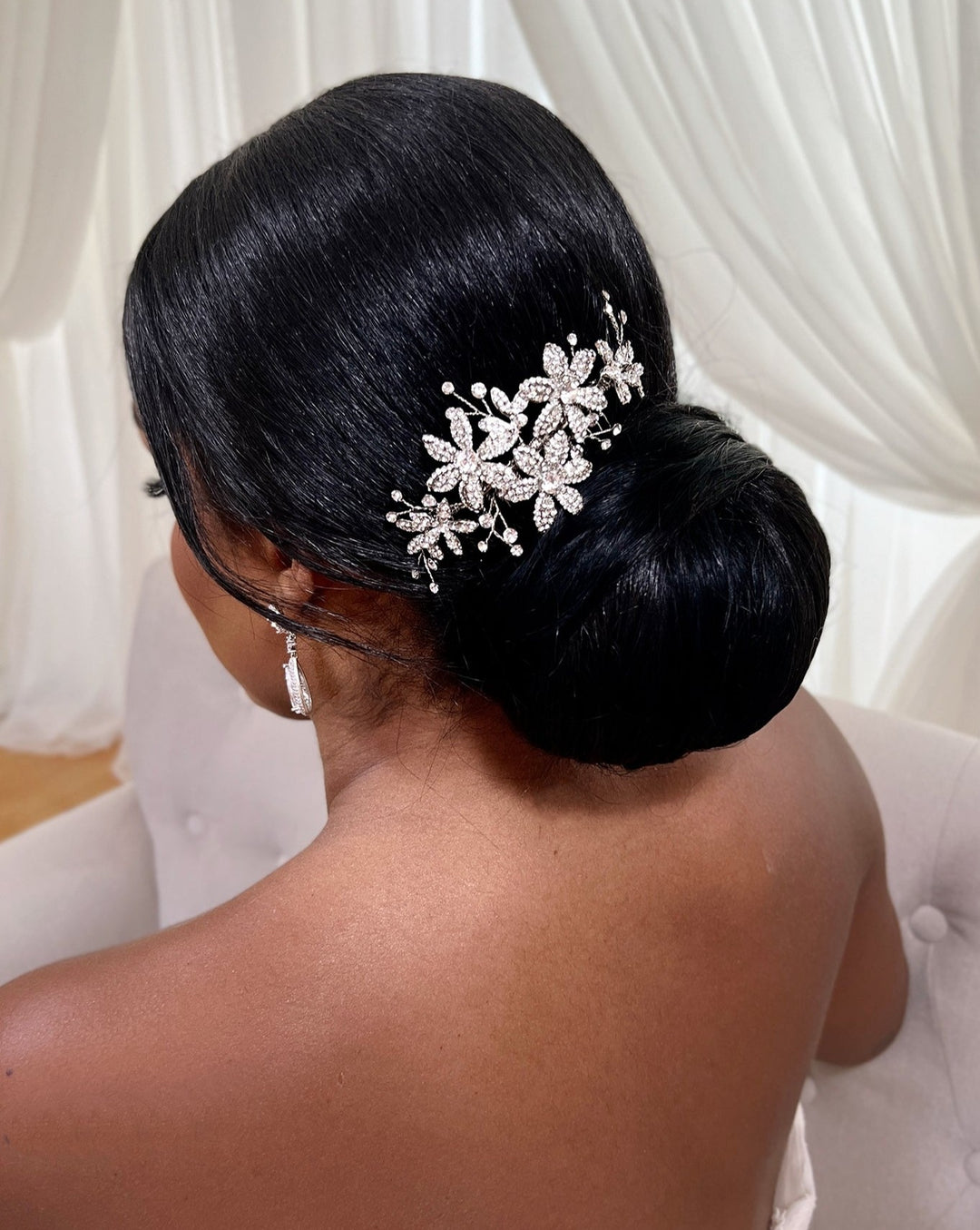 A woman with sleek, dark hair styled in a low bun adorned with the Delicate Floral Comb (style no. 242692). She is wearing elegant dangling earrings. White drapery softly blurs in the background.