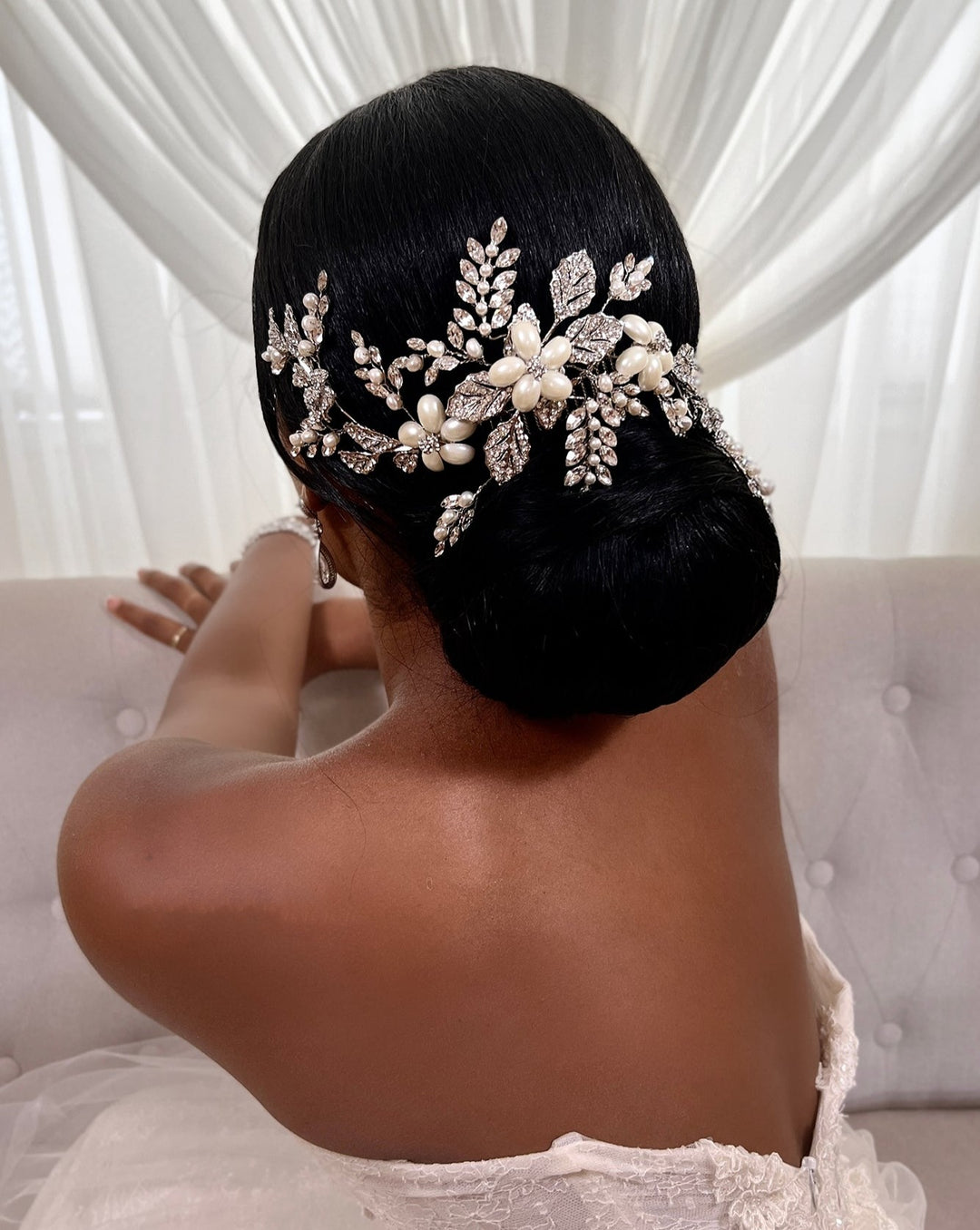 A person with an elegant updo hairstyle adorned with a Branching Pearl and Crystal Comb (style no. 80027) seated on a beige couch, wearing a strapless dress, with sheer white curtains in the background.