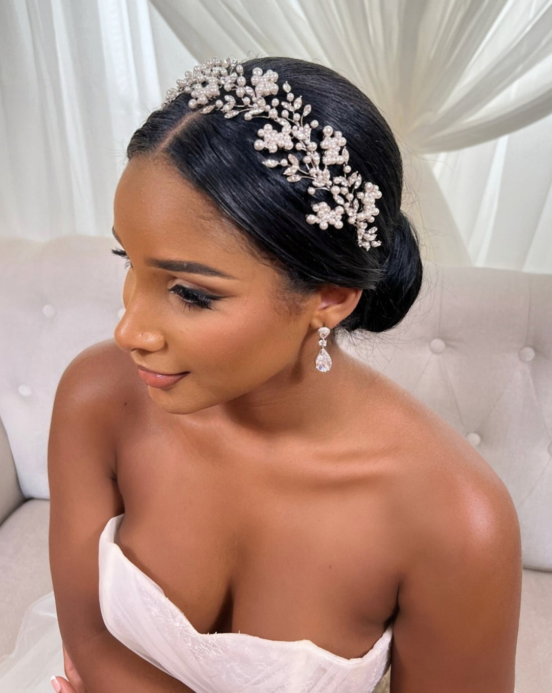 A woman in a strapless white dress sits on a light-colored sofa. Her elegant updo is adorned with the Shimmering Cluster Headband Style No. 242685, and she wears dazzling dangling earrings. Flowing white curtains in the background enhance her graceful presence.