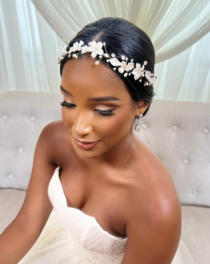 A bride in a strapless white gown, with elegant makeup, long eyelashes, and soft pink lipstick, sits gracefully on a light couch wearing the dazzling Delicate Porcelain Flower Wedding Hair Vine (style no. 242732) as her floral headpiece. Sheer white curtains enhance her timeless beauty.