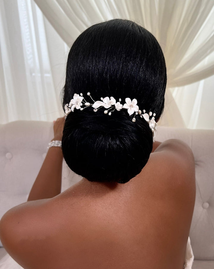Seated on a light sofa, a person with a polished black updo is adorned with the sparkling Delicate Porcelain Flower Vine, style no. 242732. The backdrop of sheer white curtains enhances the scenes soft elegance.