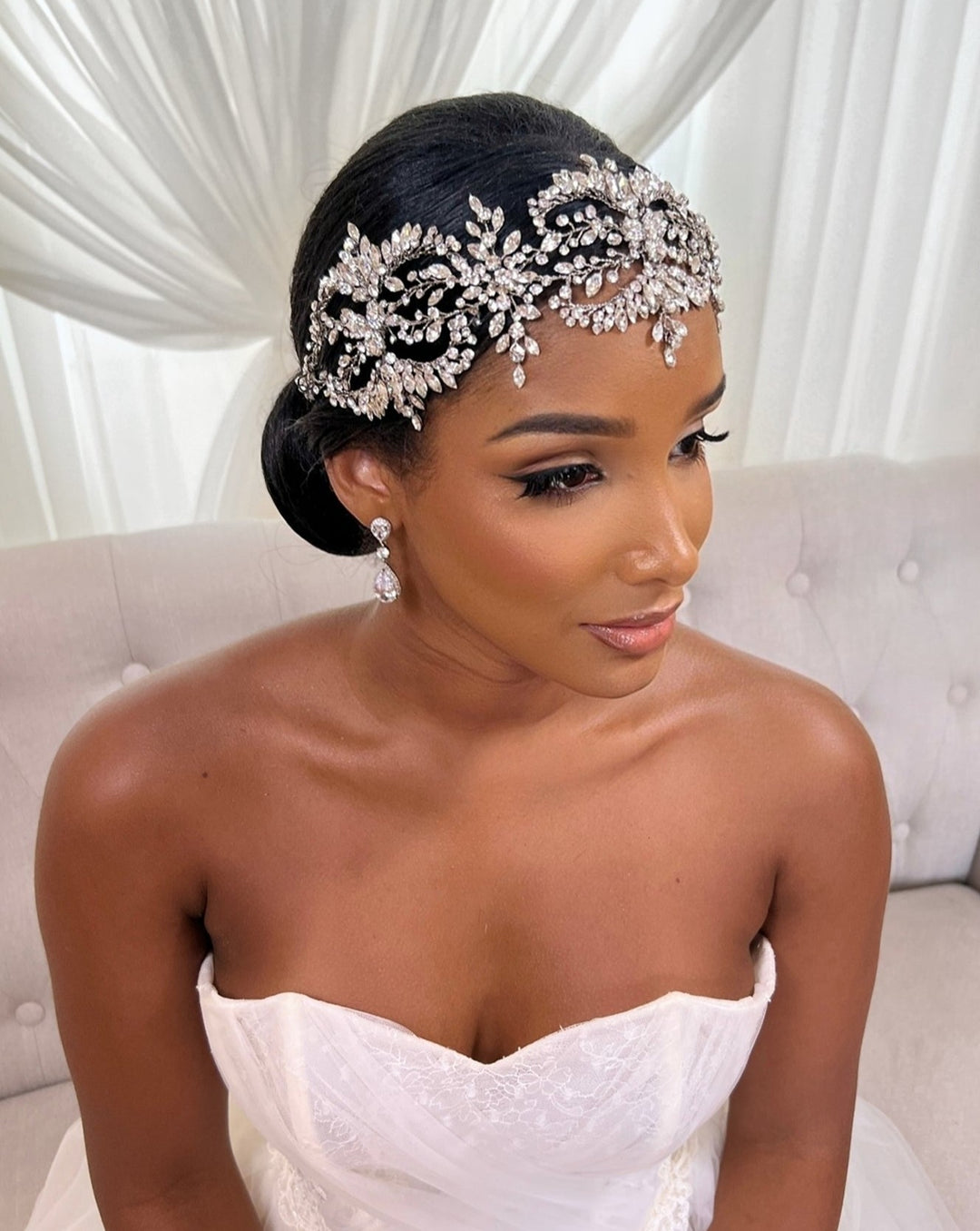 A bride in a strapless white gown sits on a sofa with her hair styled in an elegant updo adorned with the sparkling Dramatic Wreath Vine Style no. 242682 by Swarovski. Matching crystal earrings complete her glamorous look against a backdrop of white drapery.