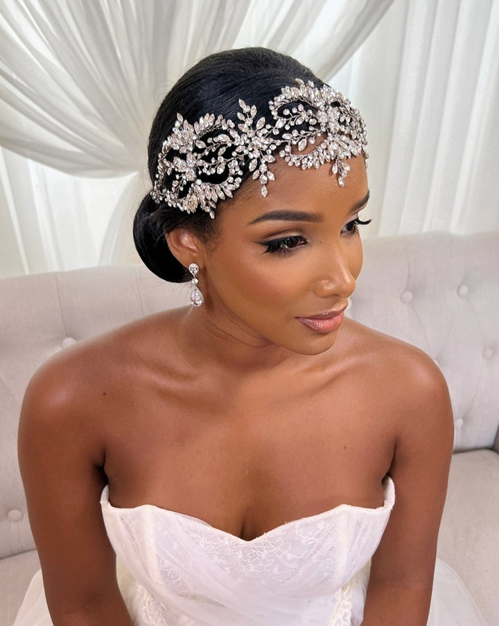 A bride in a strapless white gown sits on a sofa with her hair styled in an elegant updo adorned with the sparkling Dramatic Wreath Vine Style no. 242682 by Swarovski. Matching crystal earrings complete her glamorous look against a backdrop of white drapery.
