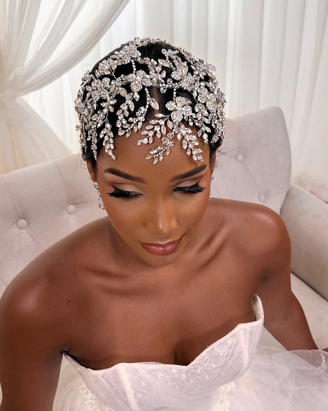 The bride wears a Dramatic Crystal Headband style no. 242695 with leaf and floral motifs, paired with matching earrings. Seated in a plush light chair, her strapless white dress is visible. She gazes downward slightly, enhanced by the elegant headband and soft makeup.