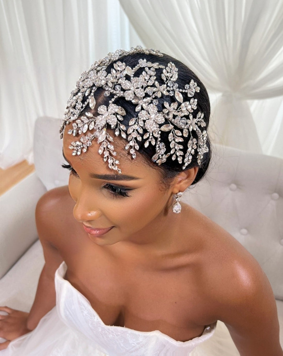 A woman in a strapless white wedding dress sits on a light-colored couch, wearing the Swarovski Dramatic Crystal Headband style no. 242695 with intricate floral and leaf crystal patterns. Sheer white curtains are in the background.