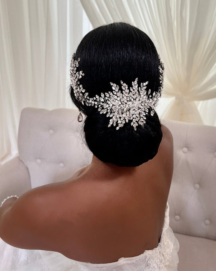 A bride with dark hair in a low bun is adorned with a Statement Swarovski Crystal Bridal Hair Vine Style no. 242551 and an off-shoulder white gown. The backdrop includes sheer white curtains and a light-colored upholstered chair.