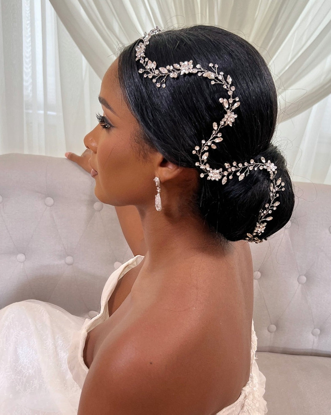 A woman in a white strapless dress with an elegant updo, adorned with the Dainty Crystal and Flower Swarovski  Crystal Hair Vine style no. 39718871. She wears dangling earrings, seated on a tufted couch and gazing to the side, while sheer curtains softly frame her silhouette.