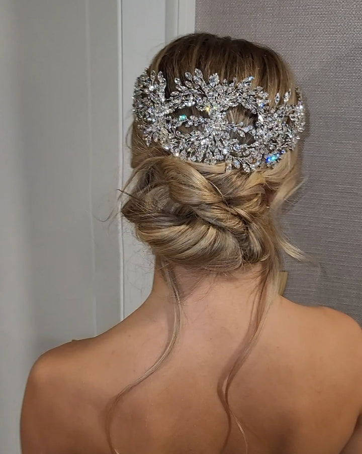 A woman with her back to the camera has blonde hair styled in a loose bun, elegantly adorned with a sparkling jeweled masquerade mask and the Couture Crystal Halo (style no. 242642), as the background softly blurs.