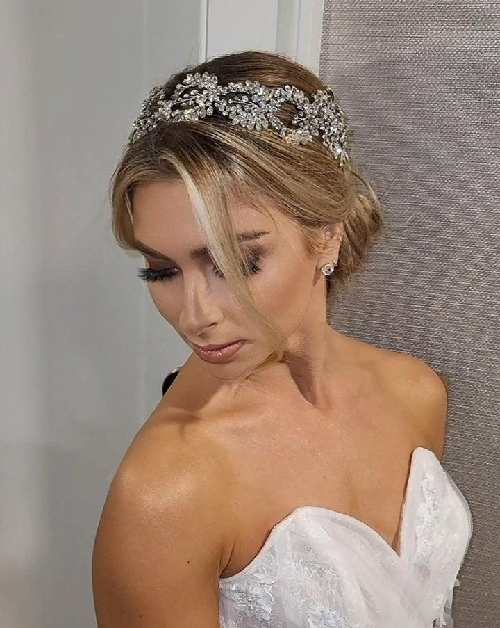 A bride in a strapless white gown and Swarovski tiara stands against a textured wall. Her hair, styled in an elegant updo, is adorned with the Couture Crystal Halo style no. 242642. She gazes downward with a serene expression.
