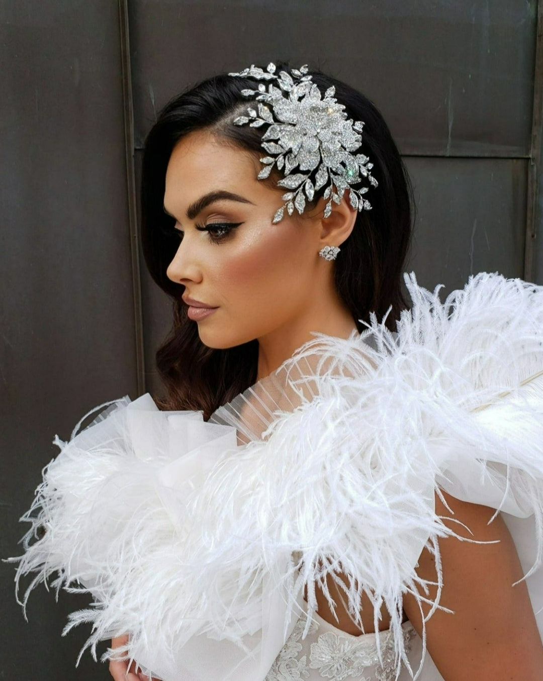 A woman with styled dark hair, wearing the Enchanting Swarovski Crystal Rose Comb style no. 242587, combined with intricate makeup and a white feathery outfit, stands in profile against a dark backdrop, exuding elegance and sophistication.