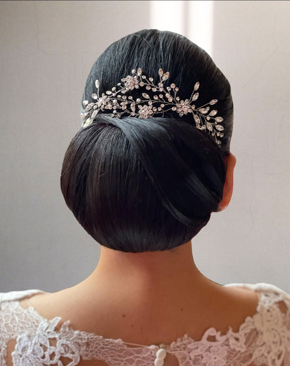 The image features the back of a woman with an elegant bun adorned with a sparkling Delicate Flowing Swarovski Crystal Vine (style no. 242464), with floral and vine motifs. Her dress showcases lace detailing on the shoulders.