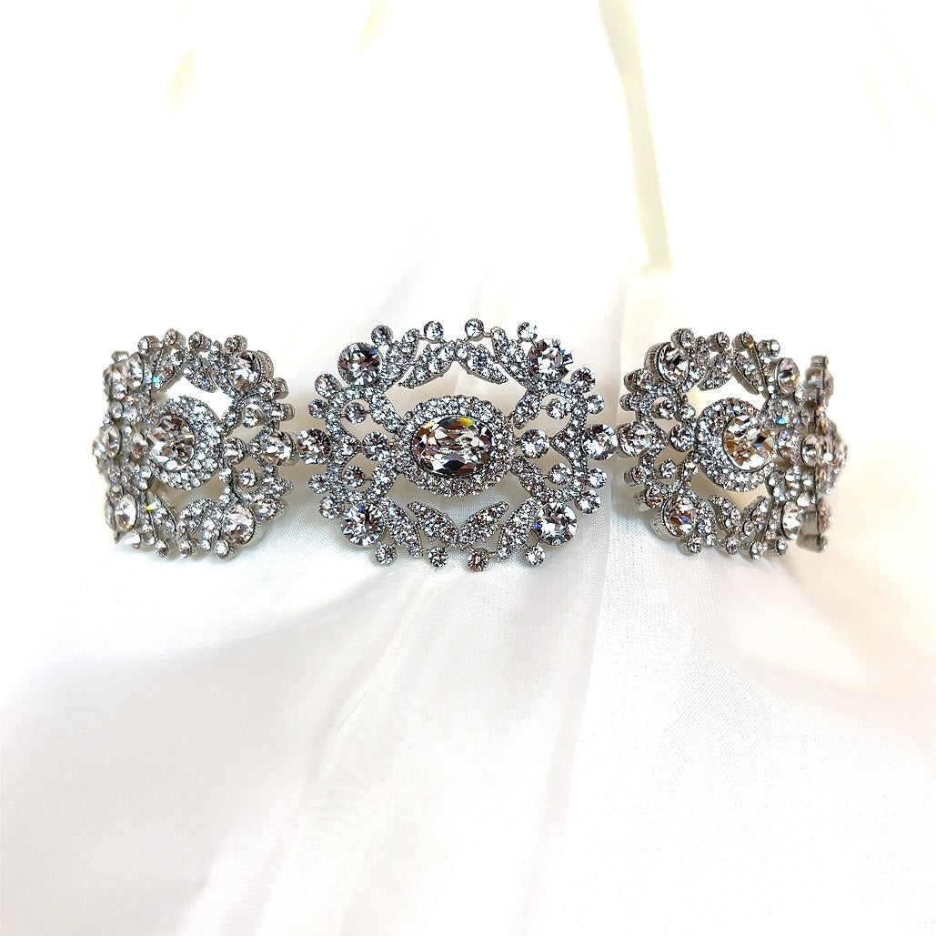 The Ornate Crystal Bridal Headband (Style no. 242737) features intricate silver floral designs and sparkling crystal accents, exuding elegance on a white fabric background, perfect for bridal wear.