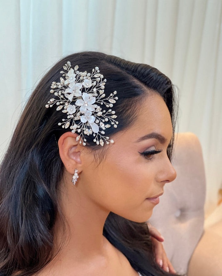 A woman with long dark hair adorns the Porcelain Flower Bouquet Comb, style no. 242722, a detailed floral Swarovski crystal hair accessories paired with matching earrings. Her elegant makeup features subtle eyeshadow and soft lipstick as she sits on a beige chair against a blurred backdrop.