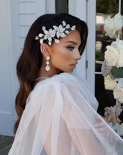 A woman in a white dress stands elegantly, her long wavy hair adorned with the Swarovski Crystal Double Flower Hair Comb (style no. 80091). She wears dangling earrings and poses gracefully in profile near a light-colored door framed by flowers.
