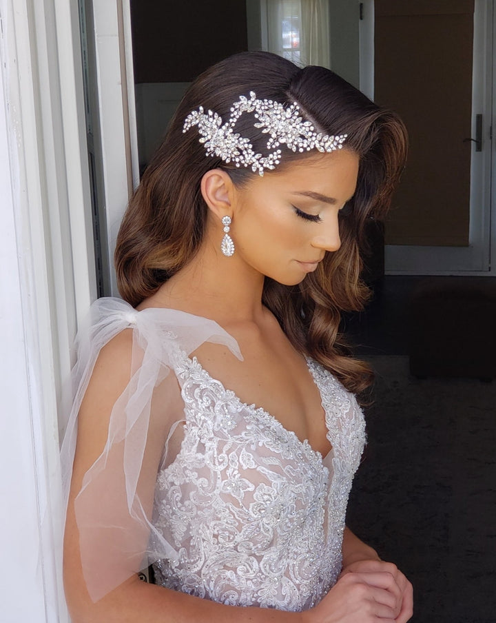 A bride stands in profile, wearing a lace wedding dress with sheer shoulders. Her long hair is styled in waves, adorned with the Flowing Cluster Hair Comb (style no. 242514) featuring genuine crystals, and matching earrings, as she calmly stands near a softly lit doorway.