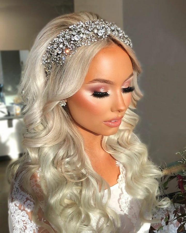 A woman with long, wavy blond hair wears the Bold and Beautiful Swarovski Crystal Bridal Headband (style no. 391022) atop her locks, adorned in earrings and a lace outfit. She gazes downward as soft lighting highlights her makeup, with bold lashes and glossy lips.