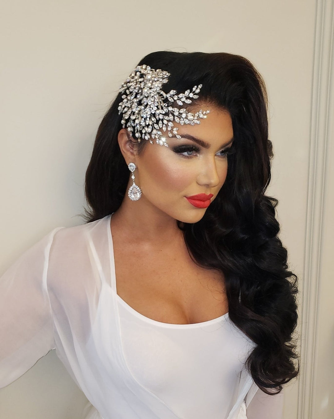 A woman with long, wavy dark hair wears a Swirling Pearl and Crystal Comb style no. 242580 and large drop earrings. Shes clad in a white outfit with sheer sleeves and bold red lipstick, striking an elegant pose against a plain background.