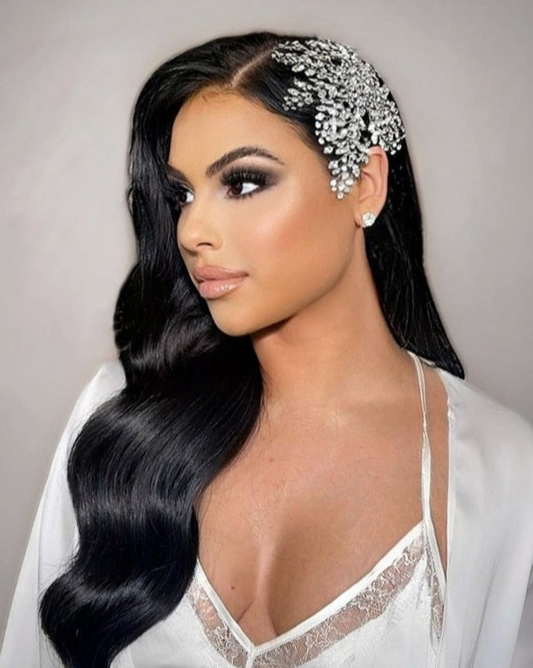 A woman with long, wavy black hair wears a Shimmering Swarovski Crystal Bridal Hair Comb style no. 242562. Shes in a white satin robe with lace details, complemented by silver earrings and neutral makeup. A soft gray backdrop enhances her sophisticated allure.