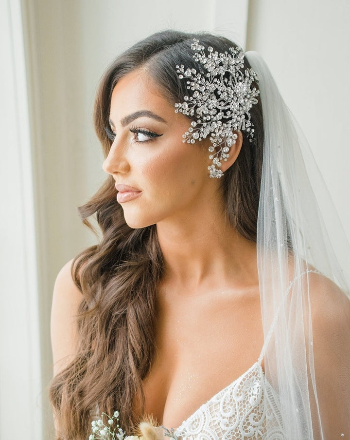 The bride, showcasing long brown hair, wears a white lace wedding dress complemented by a dazzling Swarovski crystal Snowflake Bridal Hair Comb (style no. 242431). Her veil cascades over her shoulder as she gazes pensively to the side, with soft natural lighting enhancing the scene.
