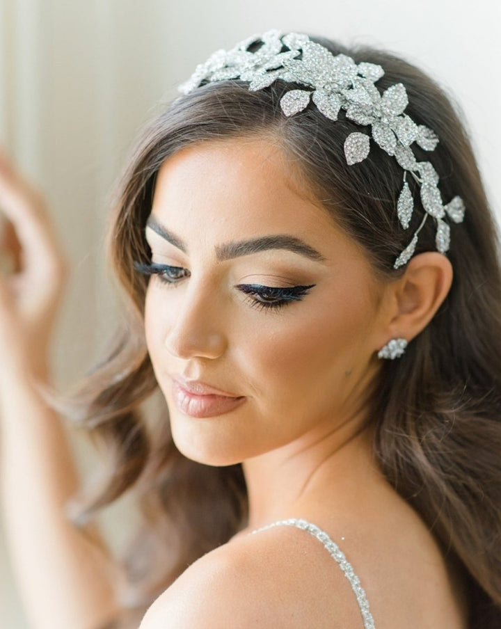 A woman with long brown hair wears a sparkling Crystal Flower Bridal Headband style no. 80070 and matching earrings. She has elegant winged eyeliner, soft pink lips, and dons a dress with thin straps, posing serenely while gazing downward.