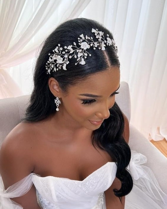A bride with long, dark hair elegantly wears a stunning  Asymmetrical Blossom Swarovski Crystal Hair Vine (style no. 242735) and matching earrings. She dons an off-shoulder white wedding gown, seated on a light chair against sheer white curtains.