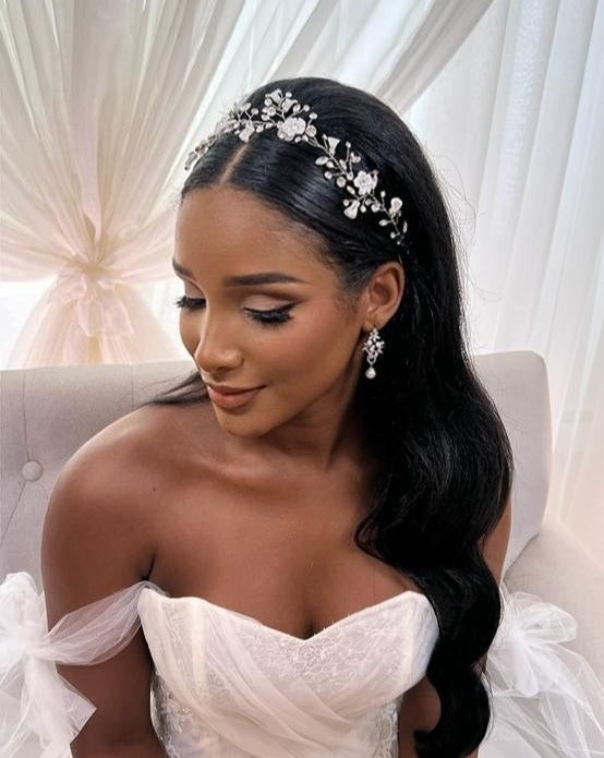 A bride in an elegant white gown sits on a light-colored sofa, serene expression, adorned with the Asymmetrical Blossom Wedding Hair Vine style no. 242735 in her long black hair. She wears pearl bridal earrings and has soft makeup. White drapes serve as the backdrop.