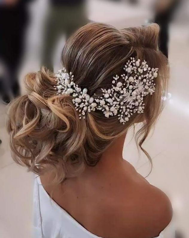 With an elegant updo a woman enhances her look with the Dramatic Asymmetrical Pearl and Swarovski Crystal Hair  Vine (style no. 242703). Shes wearing a white off-the-shoulder top against a softly blurred background, showcasing her exquisite hairstyle.