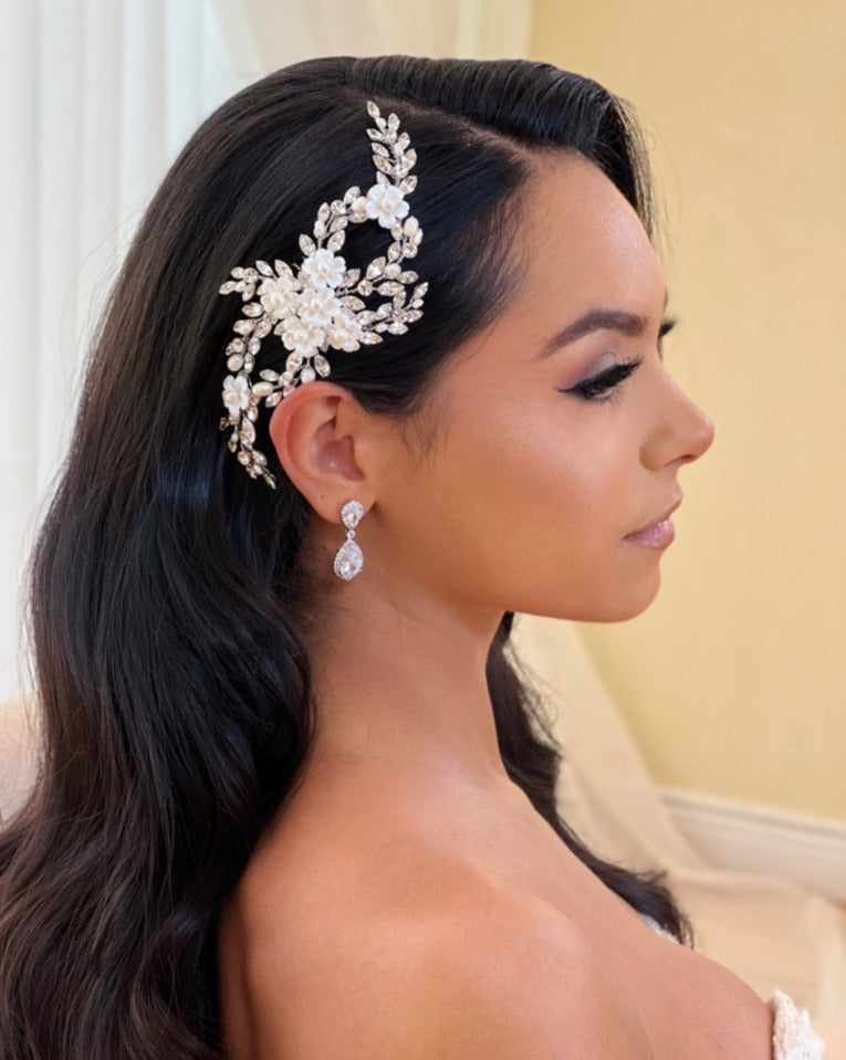 A woman with long dark hair is seen in profile, wearing the Looping Crystal and Flower Comb style no. 242725, intertwined with pearls and Swarovski crystals. Her teardrop earrings accentuate her elegant strapless outfit, all against a soft neutral backdrop.
