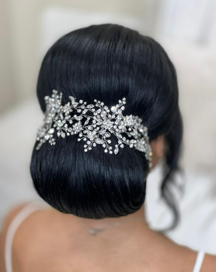 A person with long, dark hair styled in an elegant updo adorned with a large, intricate Shimmering Bridal Vine (style no. 242480) covered in Swarovski crystals. The softly blurred background highlights the stunning hairstyle.