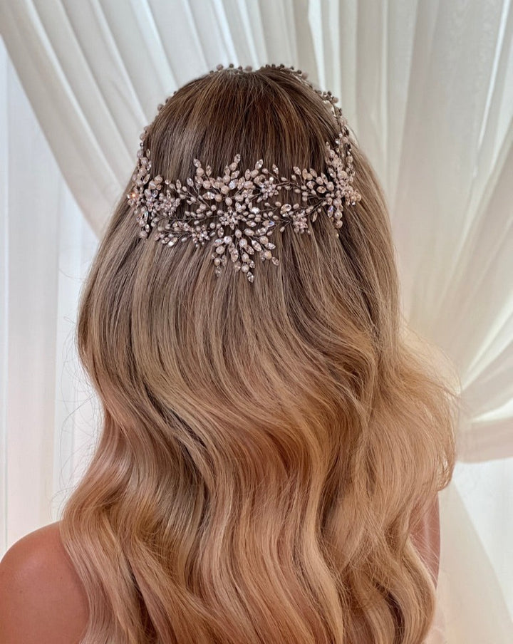 A woman with long, wavy blonde hair faces away, adorned with the Romantic Crystal and Pearl Halo (style no. 242675) by Swarovski featuring pearl-like beads and delicate branches, set against a blurred white curtain background.