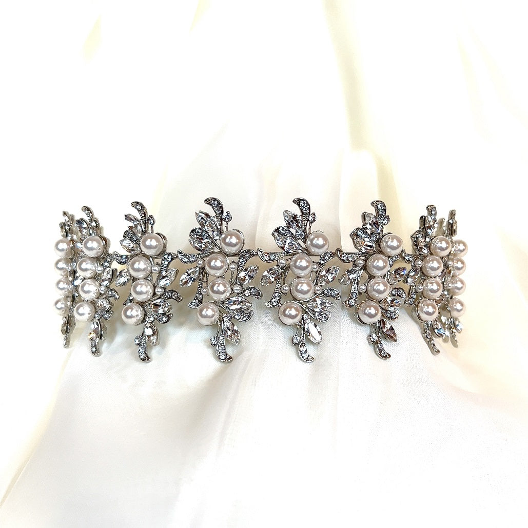 This Swarovski Luxurious Pearl and Crystal Headband (Style no.80066) features a sparkling silver design with intricate leaf patterns and multiple pearls, elegantly set against a soft, white fabric background.