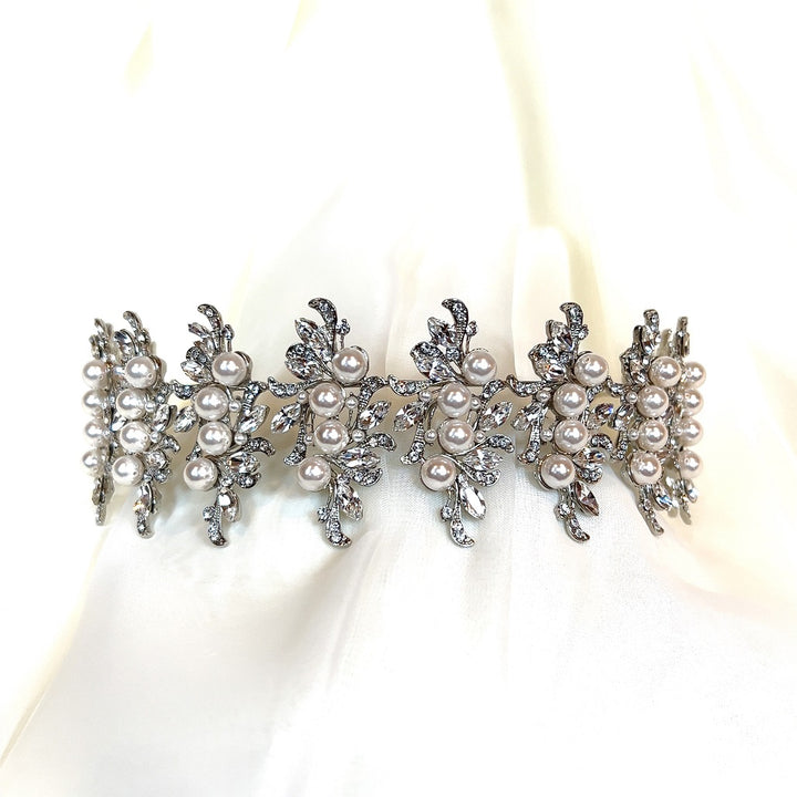 This Swarovski Luxurious Pearl and Crystal Headband (Style no.80066) features a sparkling silver design with intricate leaf patterns and multiple pearls, elegantly set against a soft, white fabric background.