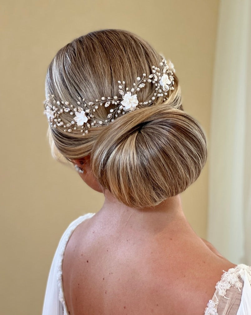 A person with blonde hair is styled in an elegant low bun, adorned by the Romantic Looping Blossom Vine (style no. 242718) featuring Swarovski crystals, white flowers, and pearl accents. They are wearing what appears to be a white wedding dress.