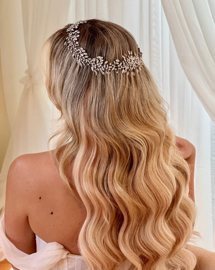 Someone with long, wavy blond hair wears the Shimmering Swarovski Crystal Bridal Halo, style no. 242542, featuring intricate floral designs. Dressed in a white off-shoulder garment, they stand before sheer white curtains.