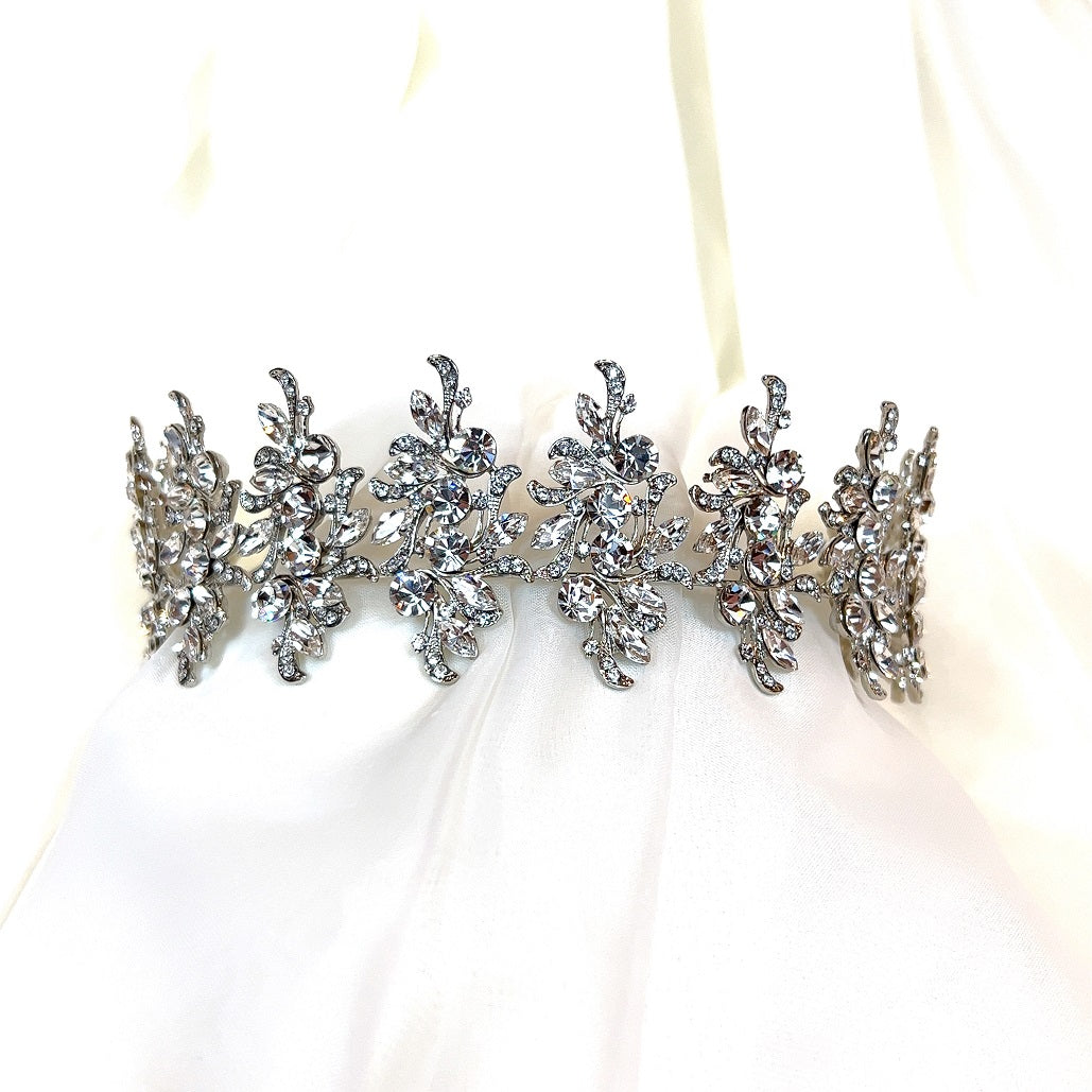 The Luxurious Pearl and Crystal Headband Style no.80066, with intricate floral designs and sparkling rhinestones, rests on a soft white fabric background. Its symmetrical leaf and petal motifs resemble a Swarovski bridal tiara, offering an elegant and ornate appearance.