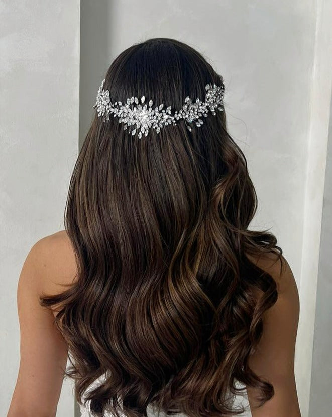 A woman with long, wavy brown hair stands with her back to the camera, wearing an Elegant Crystal Cluster Vine style no. 242517 i. The shimmering Swarovski crystals catch the light against a smooth, light gray wall.