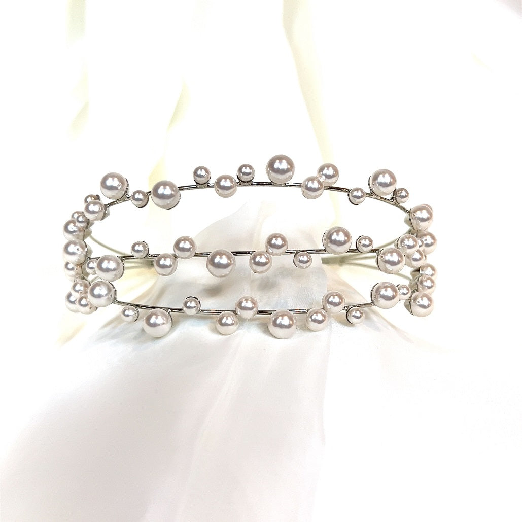 The Tiered Pearl Headband (Style no. 80096) is a Swarovski bridal tiara, featuring symmetrically placed clusters of various-sized white pearls for an elegant and delicate look, set against a soft, draped white fabric background.