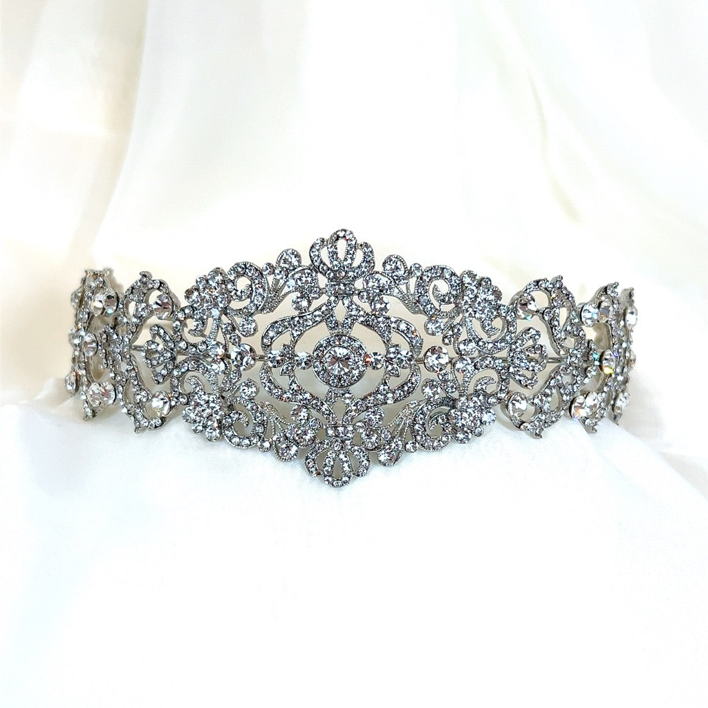 The Vintage Inspired Swarovski Crystal Headband (style no. 80107), a dazzling wedding headpiece with intricate floral and swirl patterns encrusted with crystals, is displayed on a soft, white fabric background.