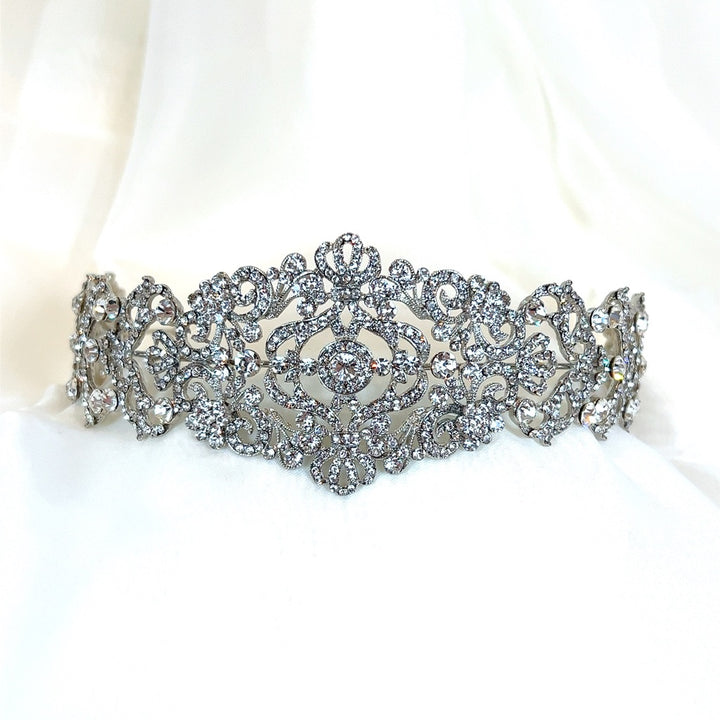 The Vintage Inspired Swarovski Crystal Headband (style no. 80107), a dazzling wedding headpiece with intricate floral and swirl patterns encrusted with crystals, is displayed on a soft, white fabric background.