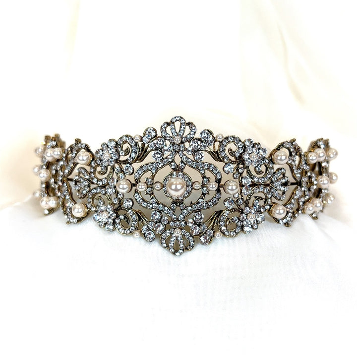 The Vintage Inspired Swarovski Crystal Headband (style no. 80107) is a glam bridal headpiece featuring intricate filigree, sparkling crystals, and delicate pearl accents. Its symmetrical design elegantly blends classic and regal elements.