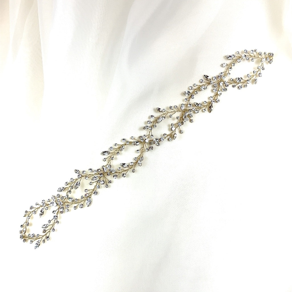 The Elegant Looping Bridal Vine, style no. 39228878, is a delicate, gold-toned headpiece with intricate leaf and vine designs adorned with small crystals, elegantly displayed on a soft white fabric background.