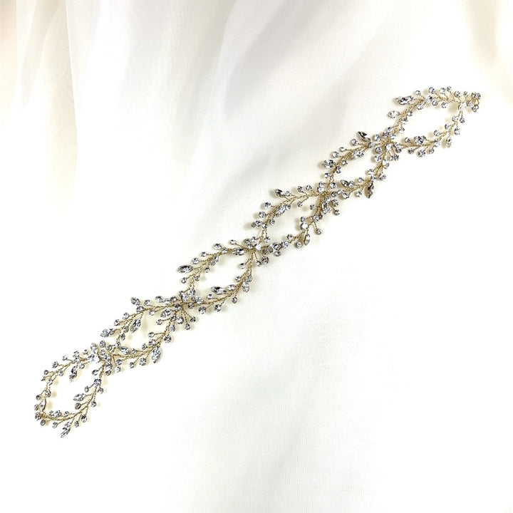 The Elegant Looping Bridal Vine, style no. 39228878, is a delicate, gold-toned headpiece with intricate leaf and vine designs adorned with small crystals, elegantly displayed on a soft white fabric background.