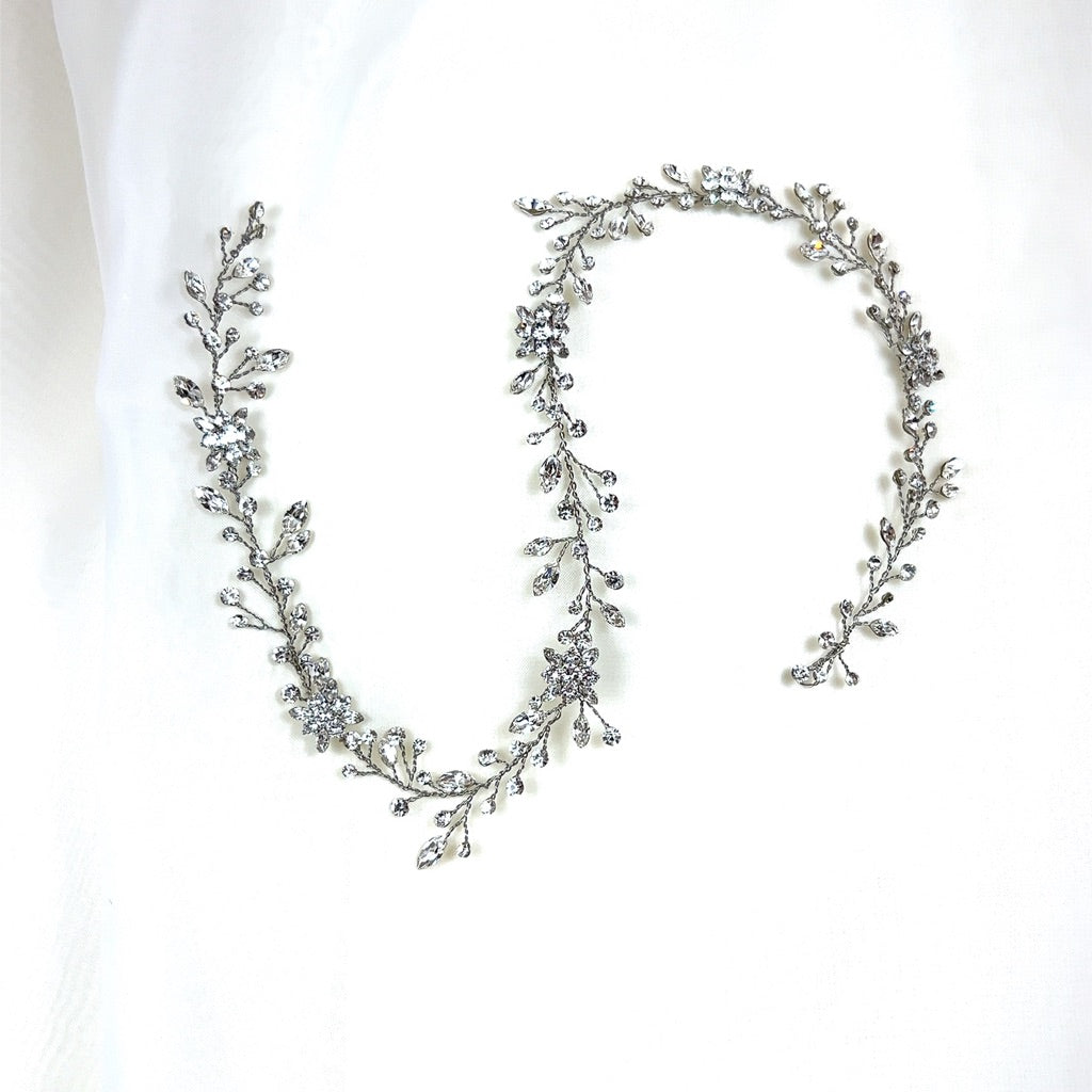 The Dainty Crystal and Flower Bridal Hair Vine (style no. 39718871) is a delicate headpiece with intricate floral and leaf designs, embellished with sparkling Swarovski crystals laid out on soft white fabric.