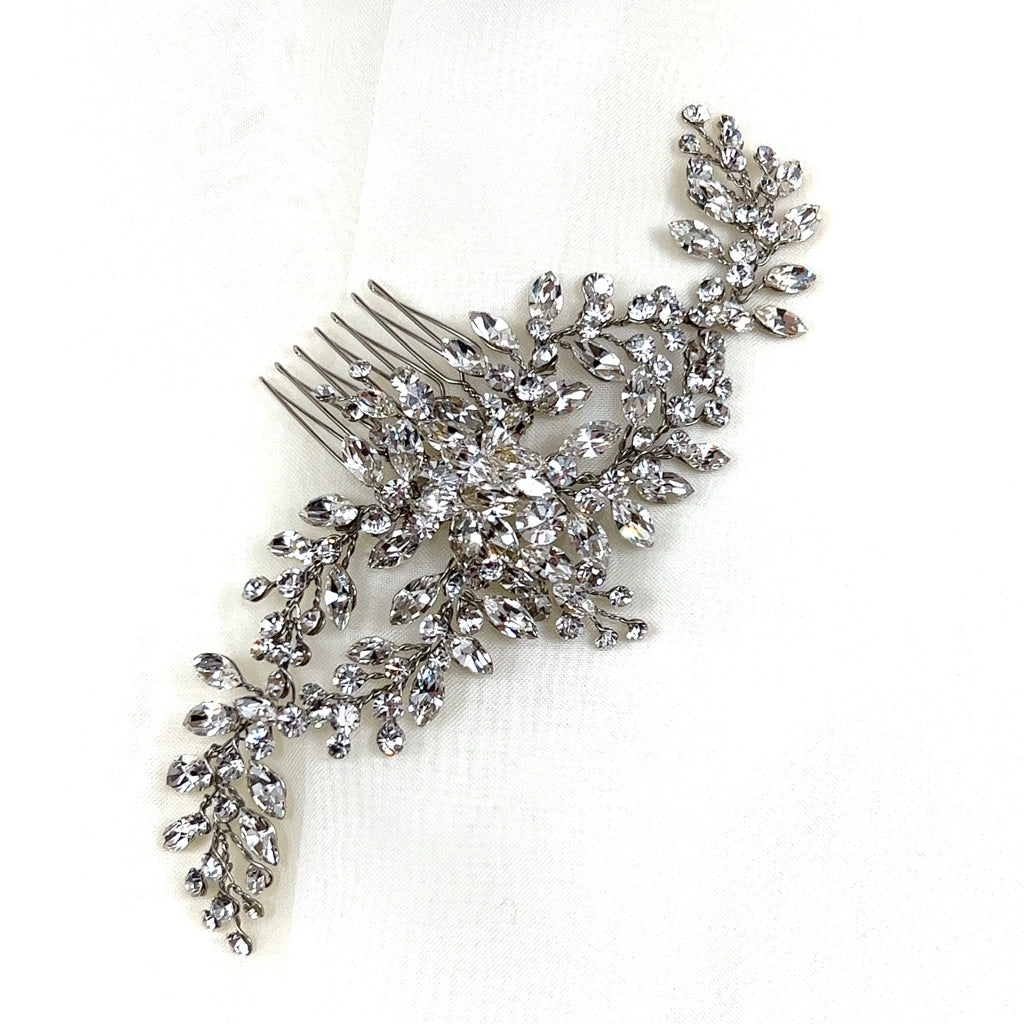 The Flowing Cluster Hair Comb (style no. 242514) is an ornate silver bridal comb featuring genuine crystals in a sparkling floral and leaf design, elegantly displayed on a soft white fabric background.