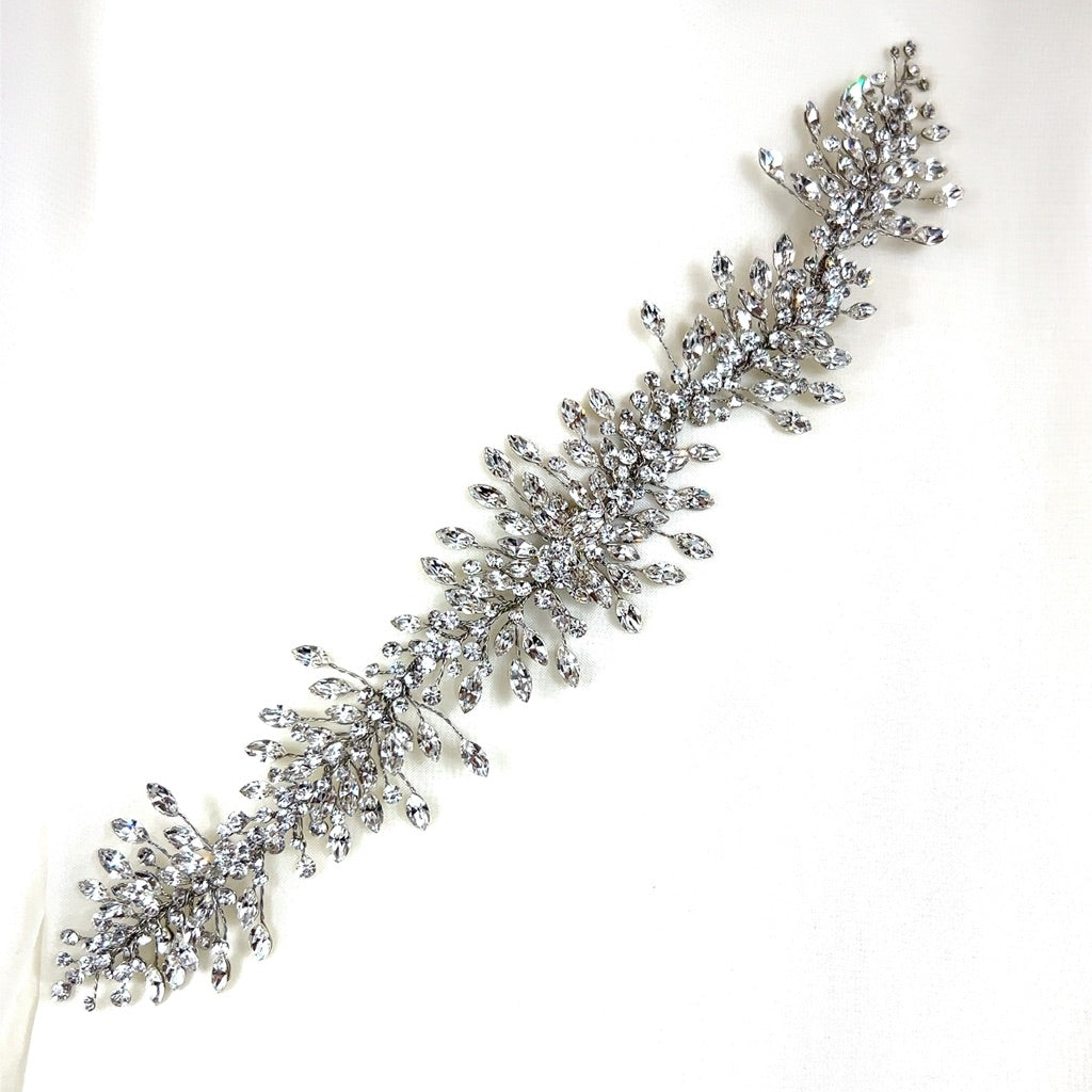 The Elegant Crystal Cluster Vine (style no. 242517) is a Swarovski bridal hair vine featuring a floral garland motif with numerous small, teardrop-shaped crystals which form leaf patterns extending from a central stem, creating an intricate look ideal for bridal ensembles.