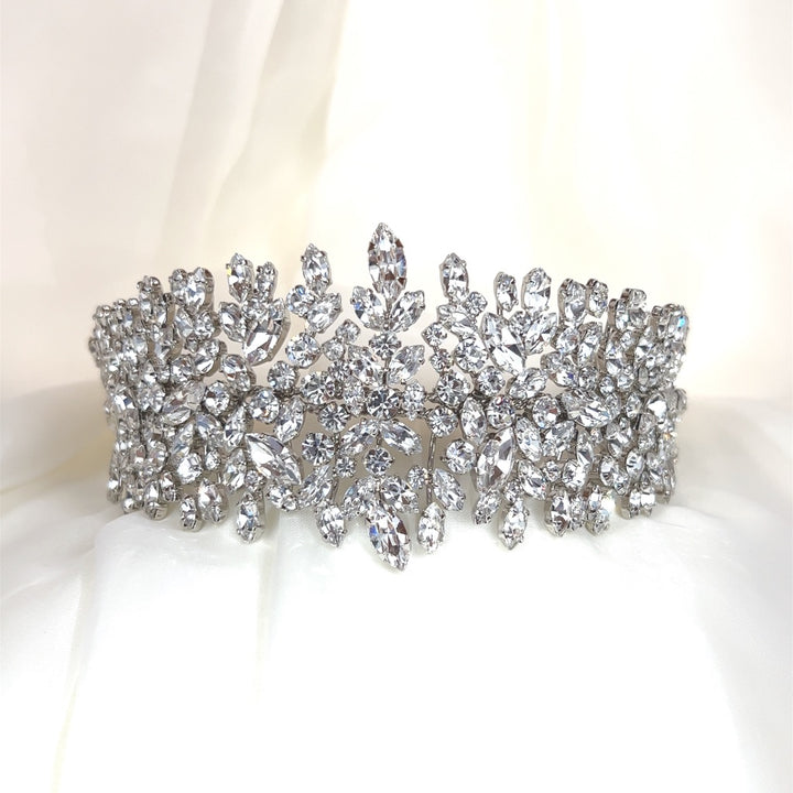 The Dazzling Crystal Headband, style no. 7900806, is a sparkling Swarovski crustal headpiece adorned with intricate clusters of dazzling leaves and floral patterns. Displayed against a soft white background, it exudes unparalleled luxury.