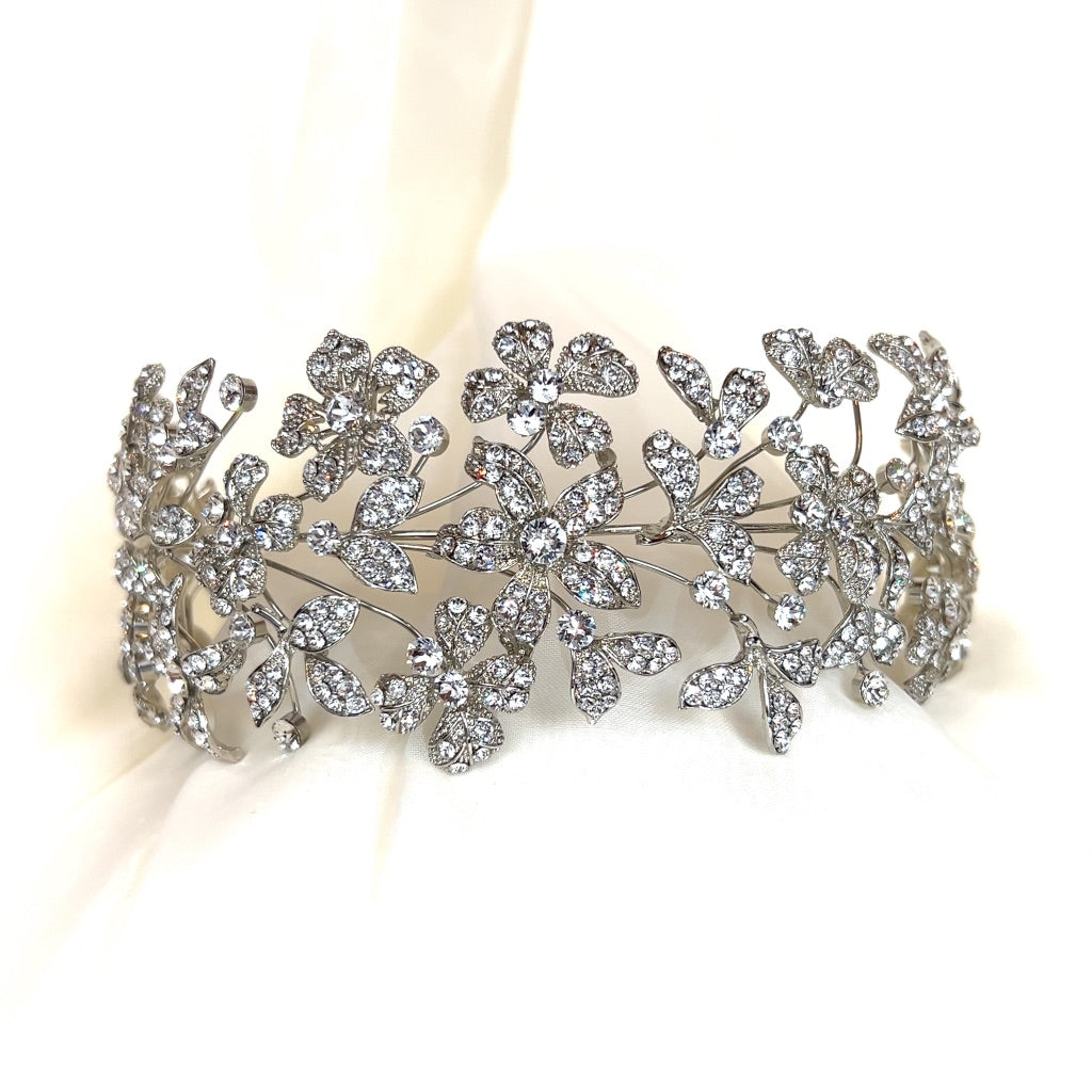 The Dazzling Crystalized Flower Headband (style no. 242678) is a sparkling bridal headpiece adorned with intricate floral patterns, each embellished with numerous Swarovski crystals. Perfect for brides, it rests elegantly on a soft white fabric background, exuding elegance and charm.