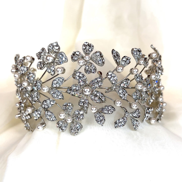 A Dazzling Crystalized Flower Bridal Headband (style no. 242678) sits on soft white fabric, adorned with intricate floral patterns of small pearls and shimmering Swarovski crystals creates an elegant and luxurious appearance.